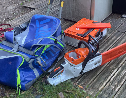 Équipement d'abattage d'arbres comprenant une tronçonneuse Stihl, un sac à dos de protection bleu et gris, une boîte à outils et des outils de sécurité sur un sol en bois à l'extérieur.
